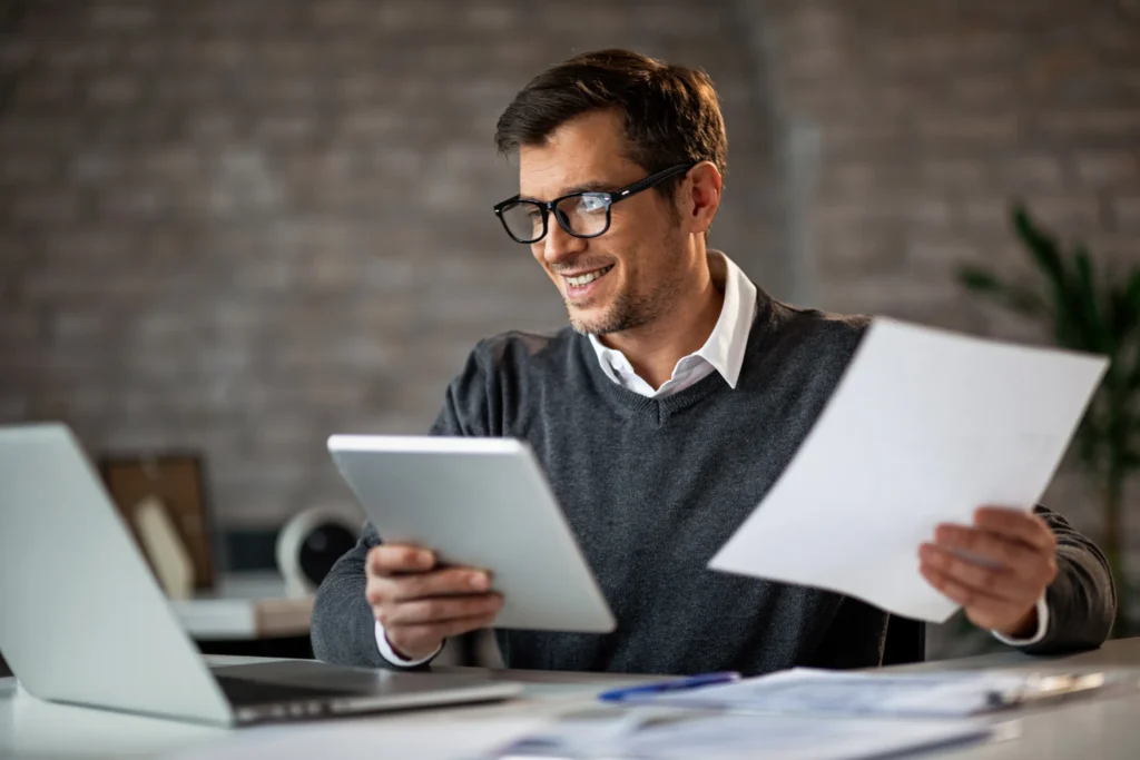 Homme souriant avec lunettes utilisant une tablette et tenant un document devant son ordinateur portable au bureau